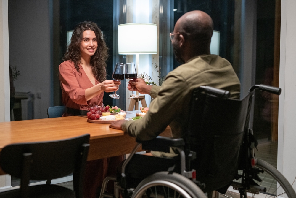 man, in wheelchair, toasting woman seated across the table with glass of red wine