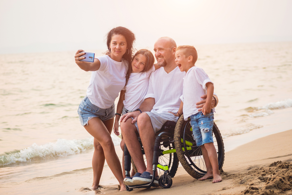 family taking a picture on the beach; mom holds camera, dad using wheelchair, two children