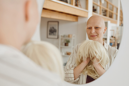 A woman with medically related hair loss looks in the mirror, holds wig.
