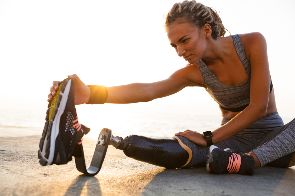 Young woman with prosthetic leg sitting on the beach doing extended leg stretches.