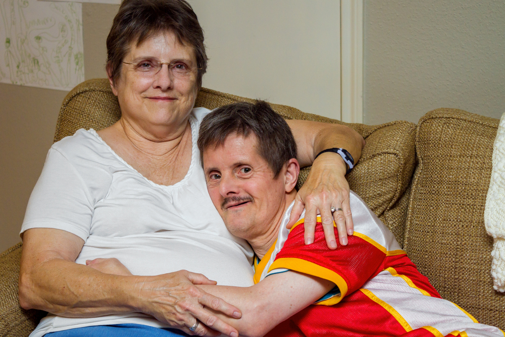 An older man with Downs Syndrome, hugs his older sister as they sit on a couch. 