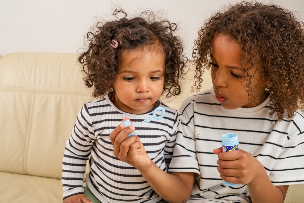 A tender sibling mixed race scene as a curly-haired girl blows bubbles for her younger sister, both dressed in matching stripes, used for National Siblings day and sisterhood. 