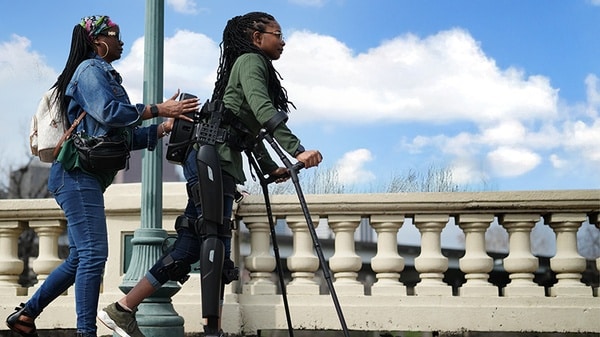 Exoskeleton device assisting woman to walk with the use of walking sticks.