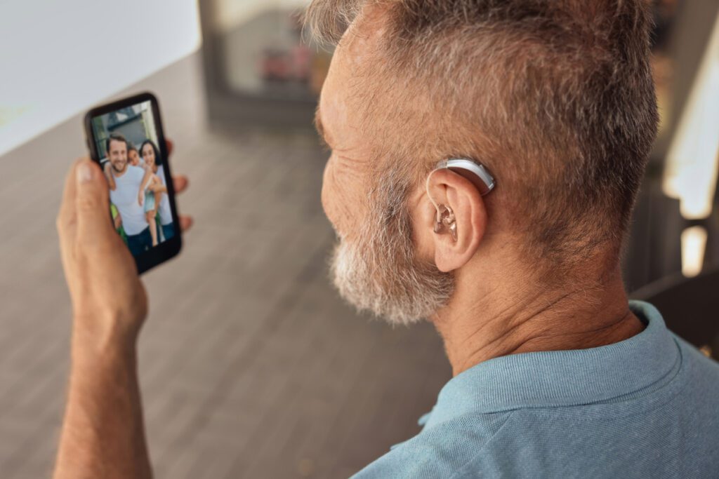 Senior man with hearing aid behind the ear communicates with his family via video communication via a smartphone. Full human life with hearing aids