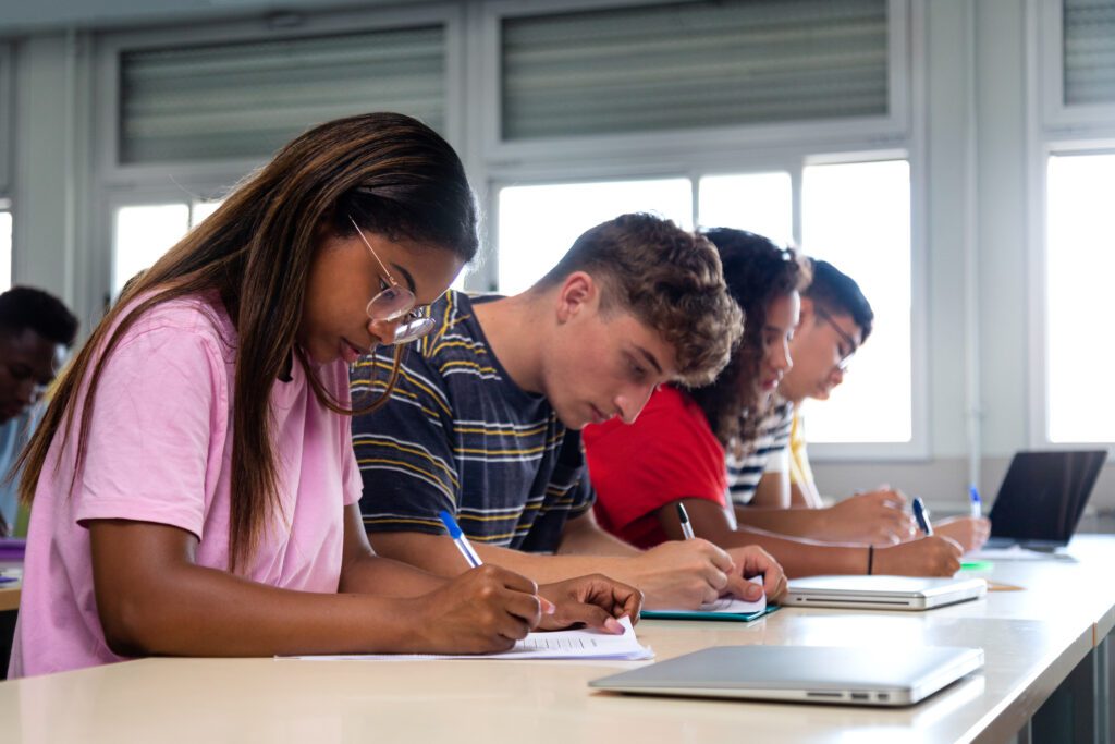 African american teen high school student writing, doing homework in class with multiracial classmates.