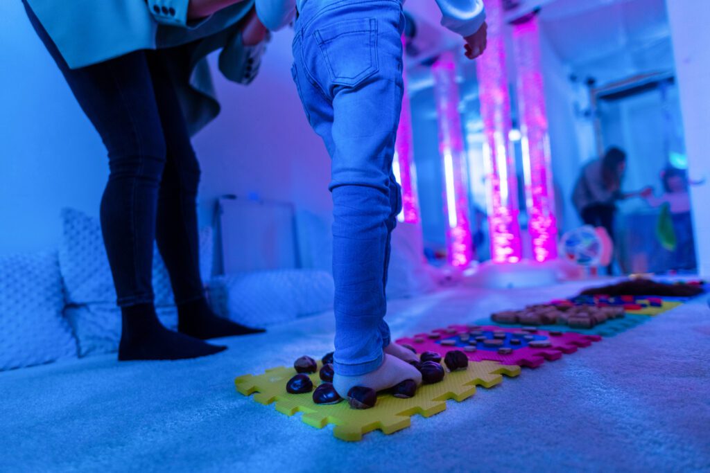 Therapist helping a young child living with cerebral palsy successfully improve her walking ability during therapy session in a sensory room, snoezelen. Texture walk, sensory exploration, motor skills.
