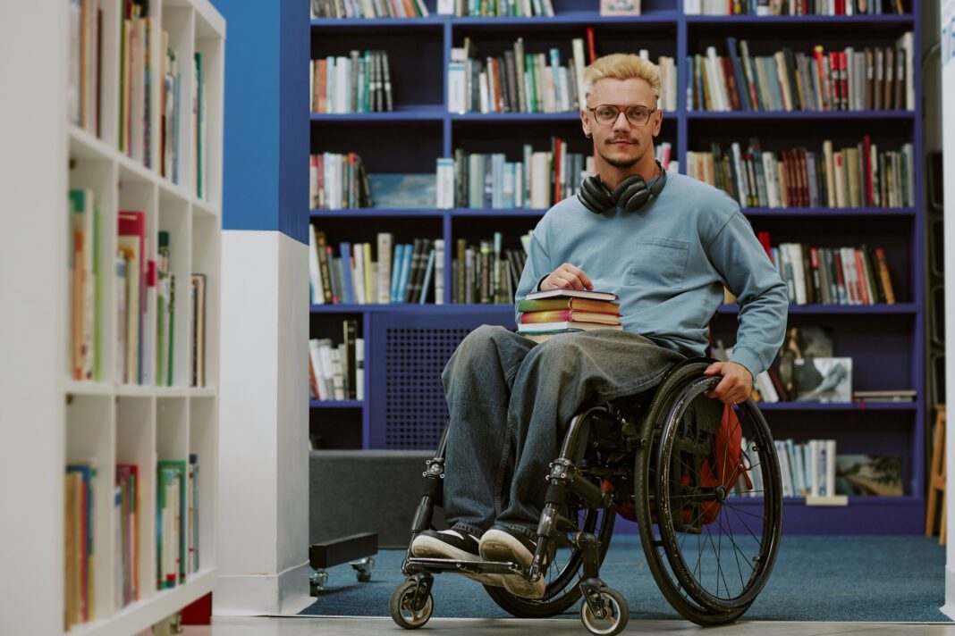 Young man sitting in wheelchair holding books while wearing headphones in library setting. He is surrounded by bookshelves filled with various titles and genres