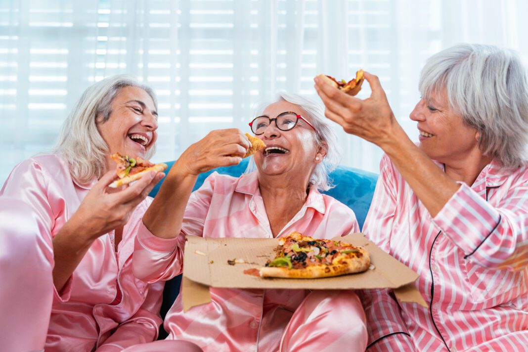 Beautiful senior women having funny pajama party at home - Group of cheerful elderly female friends having fun at home eating pizza, concepts about old people, lifestyle and quality of life
