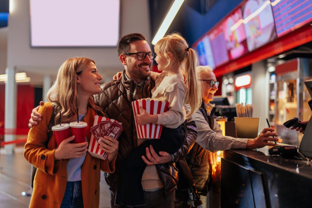 A joyful young Caucasian family with a small child is at the cinema hall, walking to the projection room with father carrying his daughter and them holding onto popcorn and drinks.